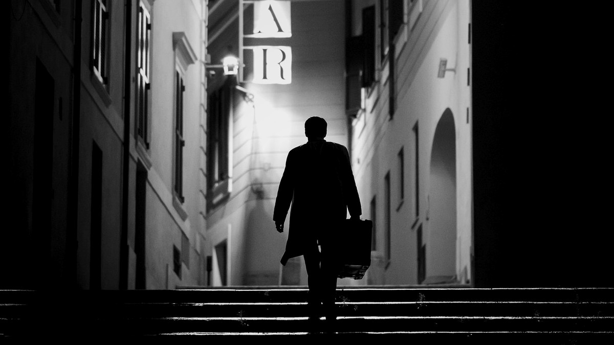 A lone silhouette walking up an Italian alley staircase under a bar sign, at night